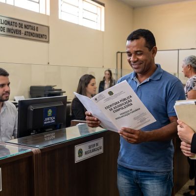 Fotografia do interior de um cartório. Em primeiro plano, um homem sorridente de camisa azul e sua advogada de terno escuro leem juntos um documento onde está escrito "ESCRITURA PÚBLICA DE ADJUDICAÇÃO COMPULSÓRIA - PROCESSO DE REGULARIZAÇÃO FUNDIÁRIA". Ao fundo, há um atendente no guichê e placas indicando "TABELIONATO DE NOTAS E REGISTRO DE IMÓVEIS - ATENDIMENTO", "SETOR DE REGULARIZAÇÃO" e "ENTREGA DE DOCUMENTOS".
